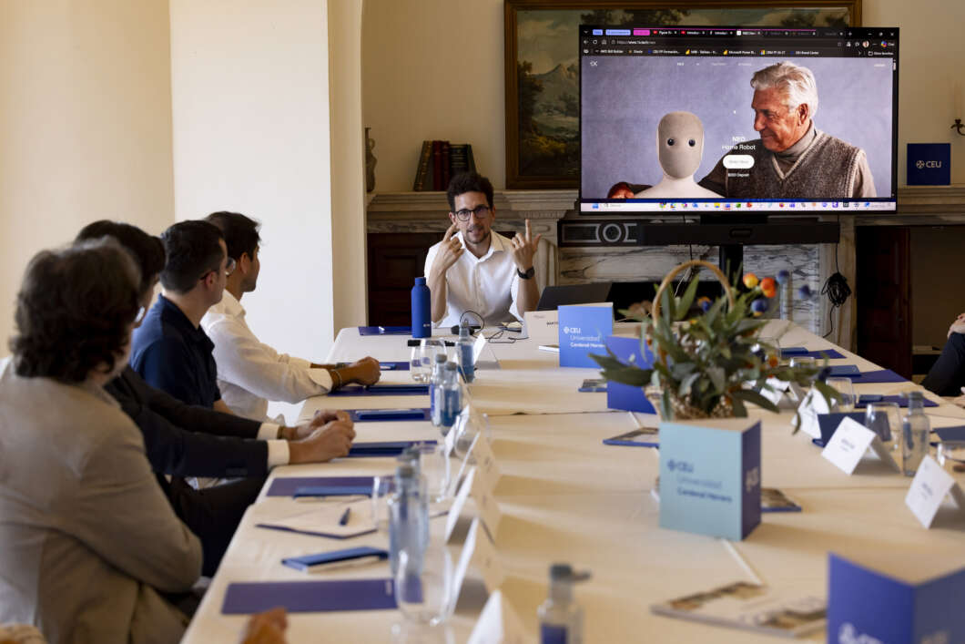 Presenter explaining to a group around a long conference table, with a large screen showing a robot and an older man in the background.