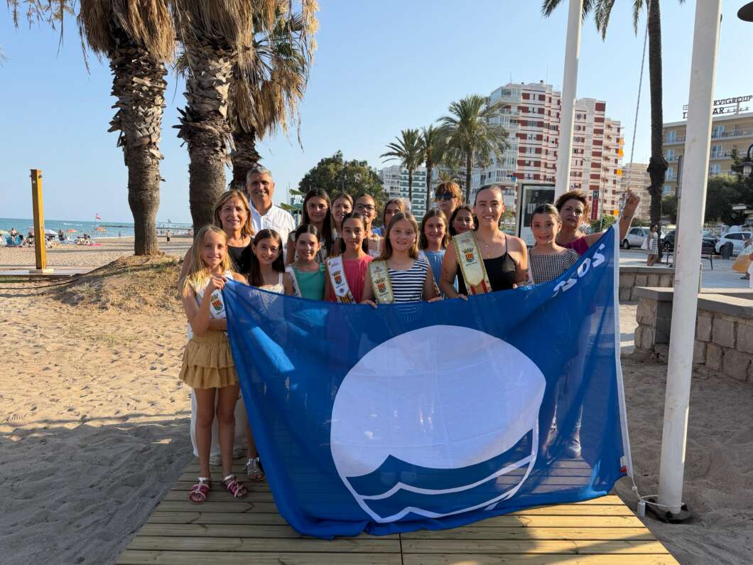 Group of smiling children and a few adults posing on a beach, holding a large blue flag with a white sun/wave emblem.