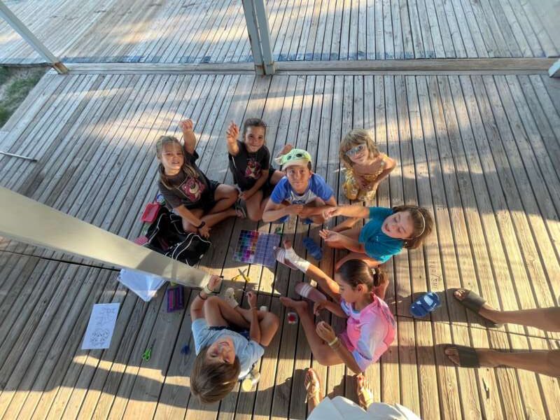 Group of children sitting on a wooden deck, looking up at the camera, with craft supplies scattered between them outdoors.