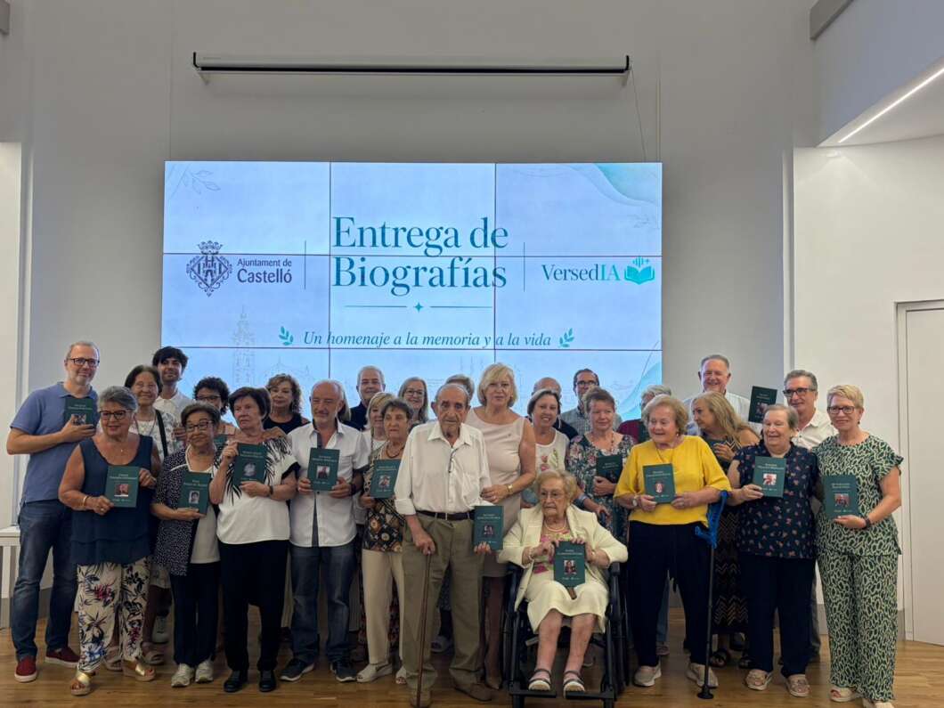 Group of about 25 people posing together, each holding teal booklets, in front of a large screen that reads 'Entrega de Biografías' (Biography Presentation).