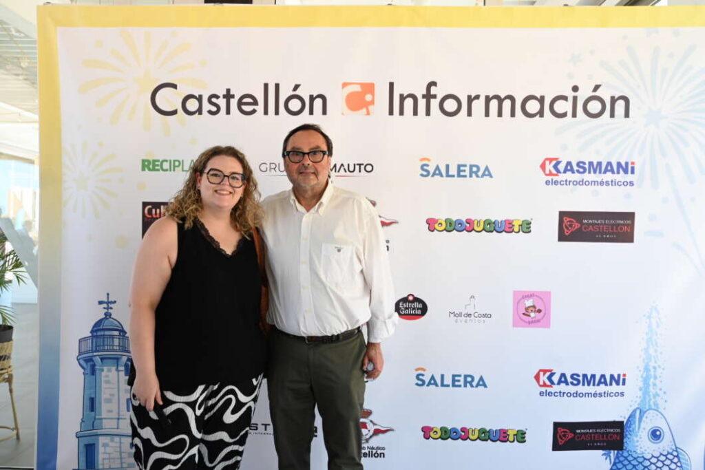 Two people stand and smile in front of a Castellón Información backdrop with sponsor logos at an event venue.