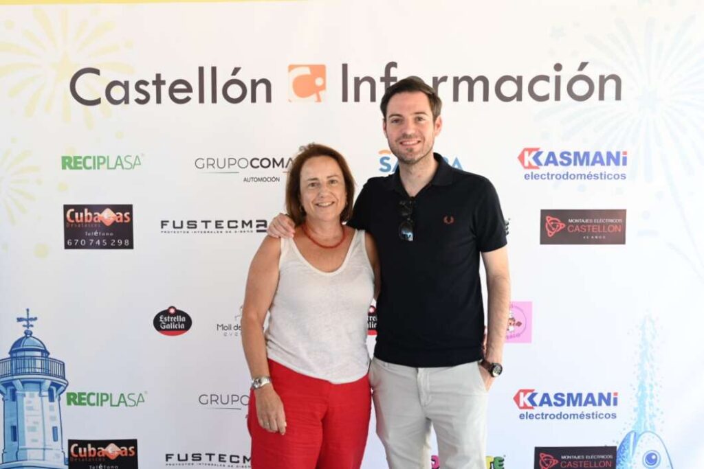 Two people smiling and posing together at a Castellón Información event backdrop. A woman in white top and red pants stands beside a man in a black polo and light pants, arms around each other.