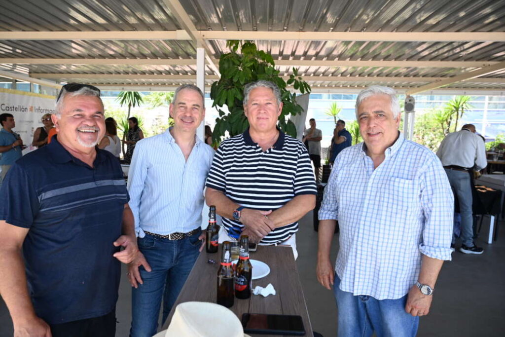 Four middle-aged men smile and pose around a tall table with beer bottles at an outdoor social event under a covered structure.