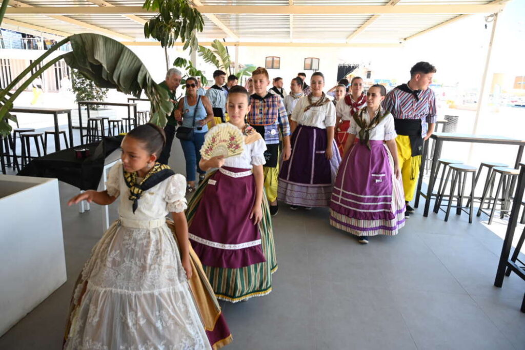 Group of people in traditional colorful dresses walking in a bright room, one girl holds a fan.