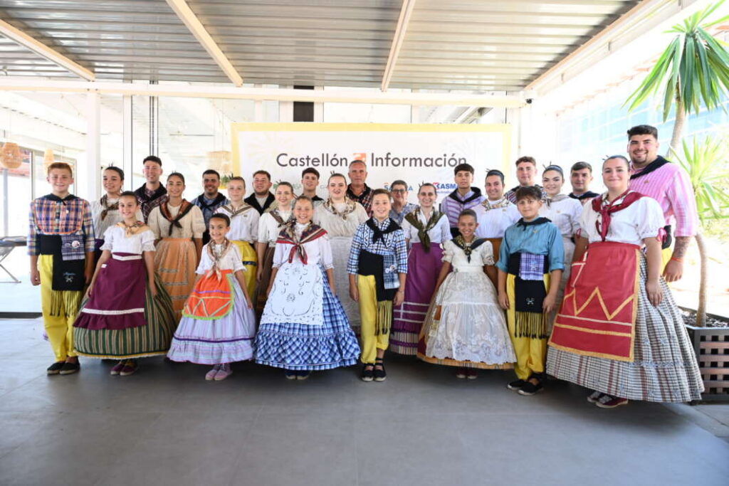 Group of people in colorful traditional regional costumes posing indoors in front of a Castellón Información banner.