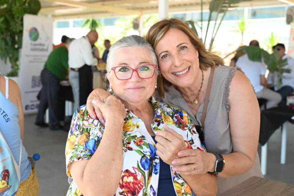 Two women pose and smile for the camera at a social event, one wearing a colorful floral shirt and red glasses, the other wrapping an arm around her shoulder in a friendly hug.
