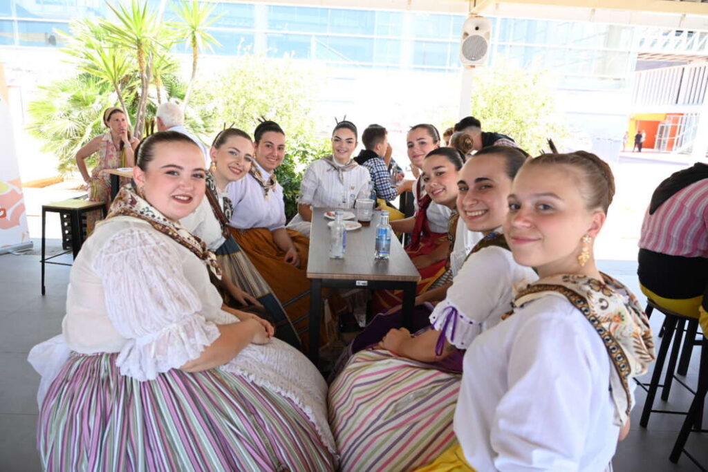 Group of women in traditional folk dresses sitting around a table outdoors, smiling at the camera.
