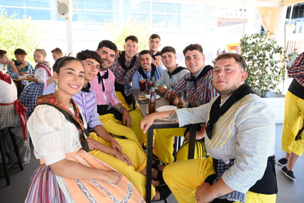 Group of people in traditional clothing sitting around a high table with drinks, smiling at a social gathering.
