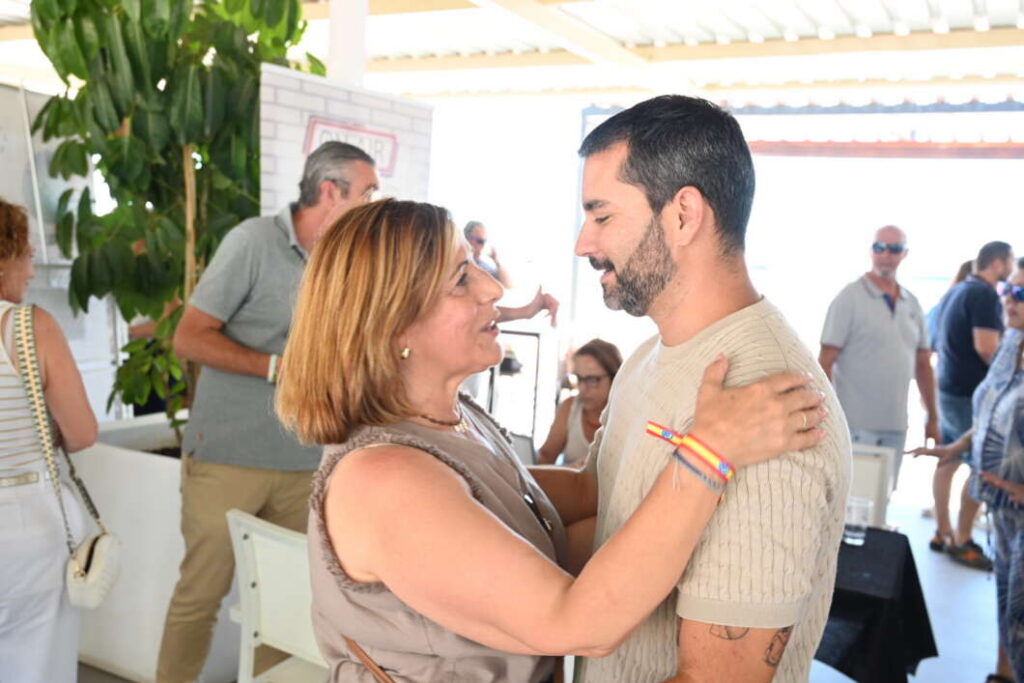 Two people embrace and smile at a social event, likely a hug between a woman and a man in a casual setting.