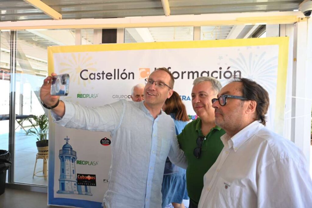 Three men pose for a selfie in front of a Castellón Información banner at a sponsored event.