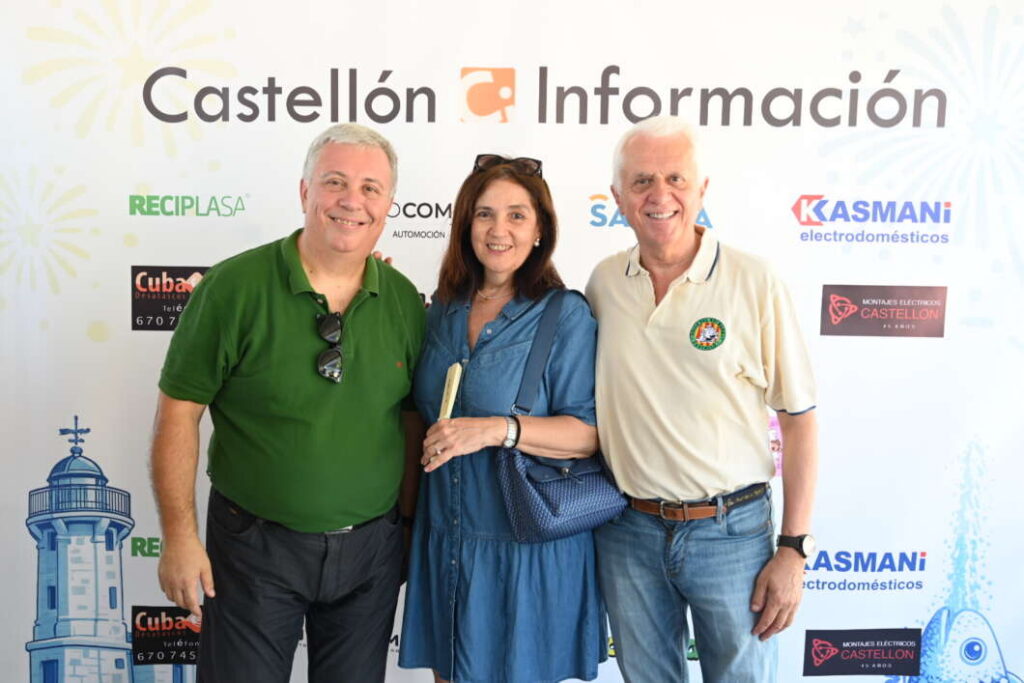 Three smiling people pose for a photo at a Castellón Información backdrop with sponsor logos behind them (left man in green polo, woman center in blue dress, right man in beige polo).