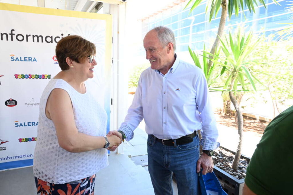 Two adults shake hands and smile at an information booth, with logos on a banner behind them and sunny plants nearby.