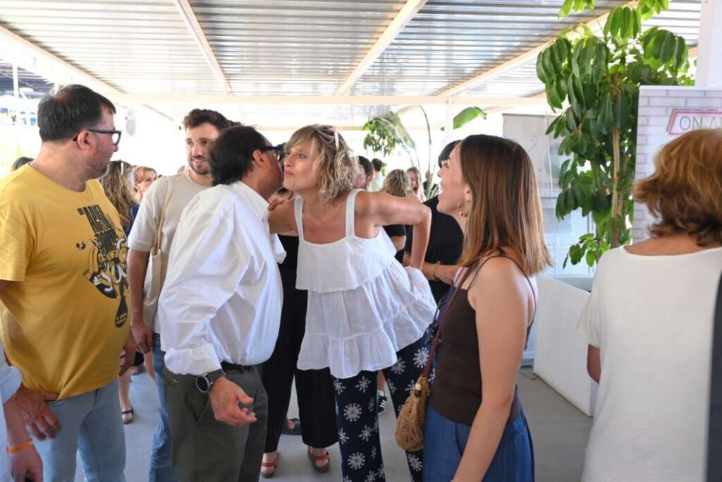 Group of adults at a bright indoor social event, chatting and greeting with smiles in a sunlit space.