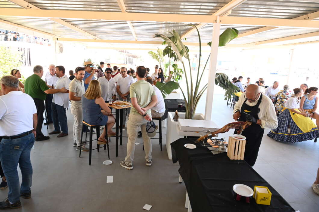 A group of people mingling at a casual outdoor event with a chef carving meat at a black-draped table on the right.