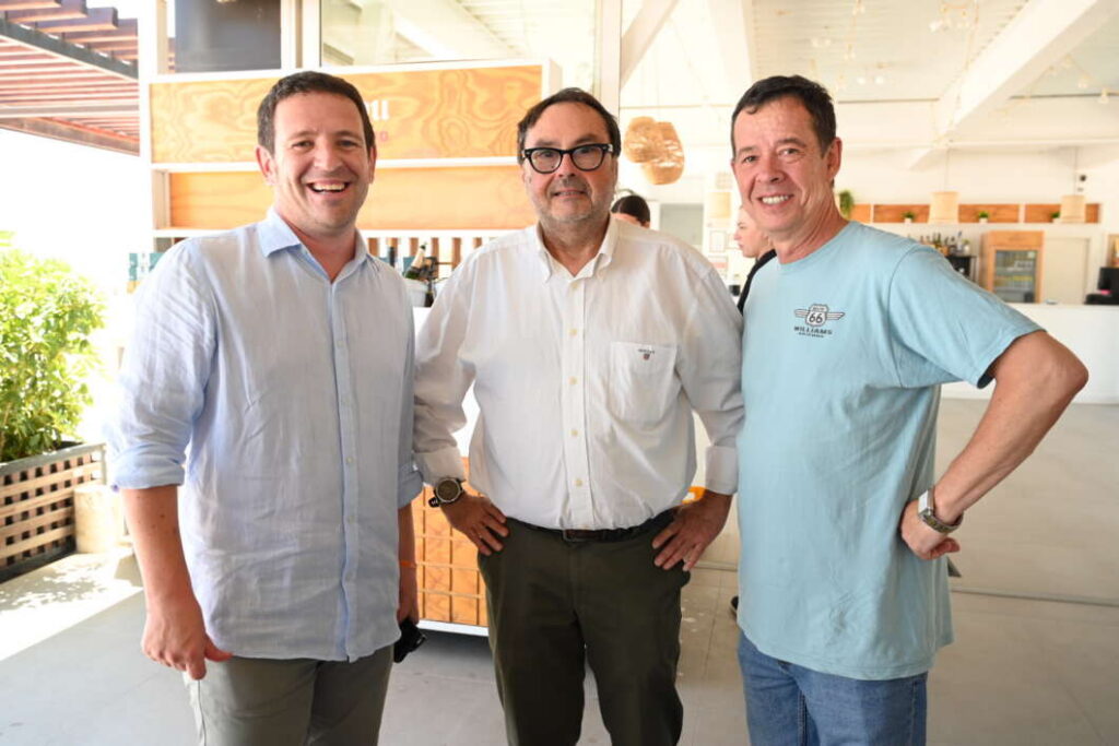 Three men stand together indoors, smiling for the camera in a casual setting.