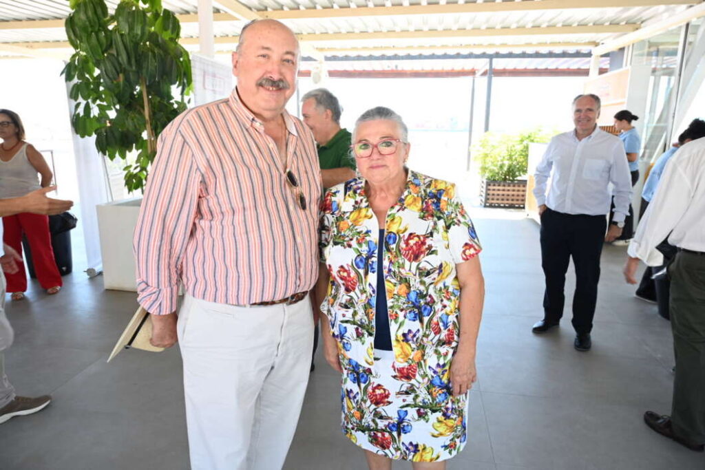 Two smiling older adults pose together at a bright terrace event; man in striped shirt and light pants, woman in a colorful floral dress.