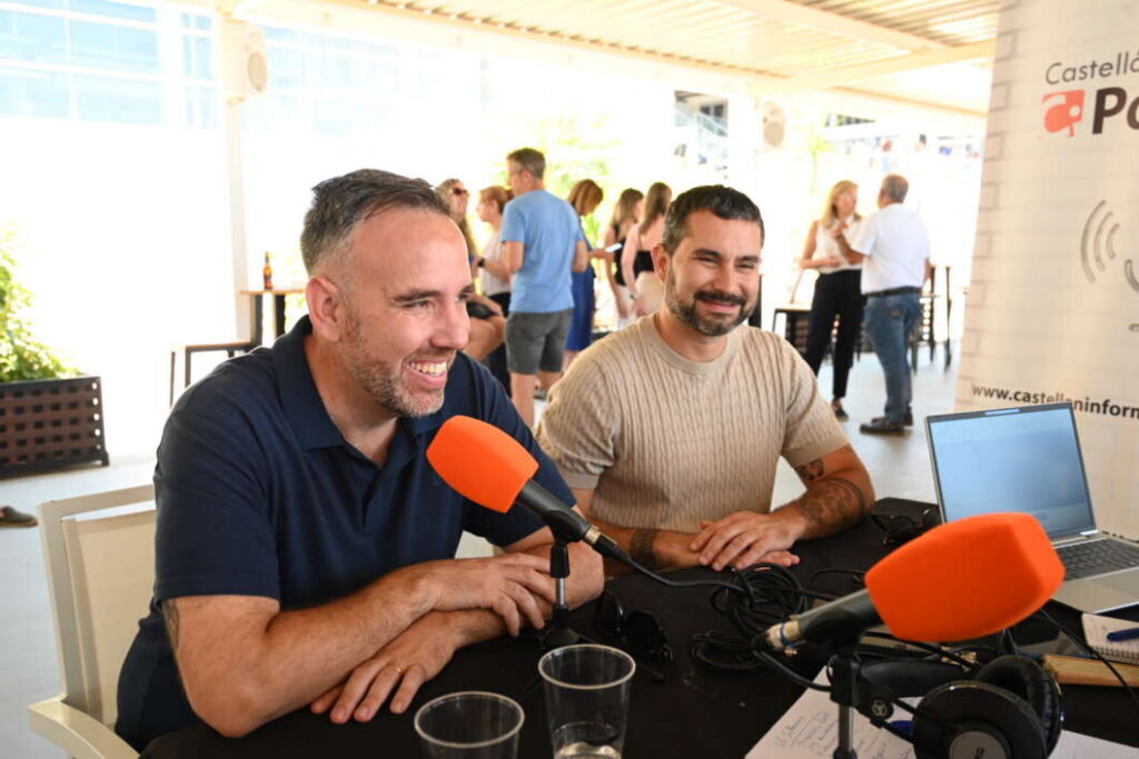 Two men sit at a table during a live interview with orange microphones and a laptop nearby.