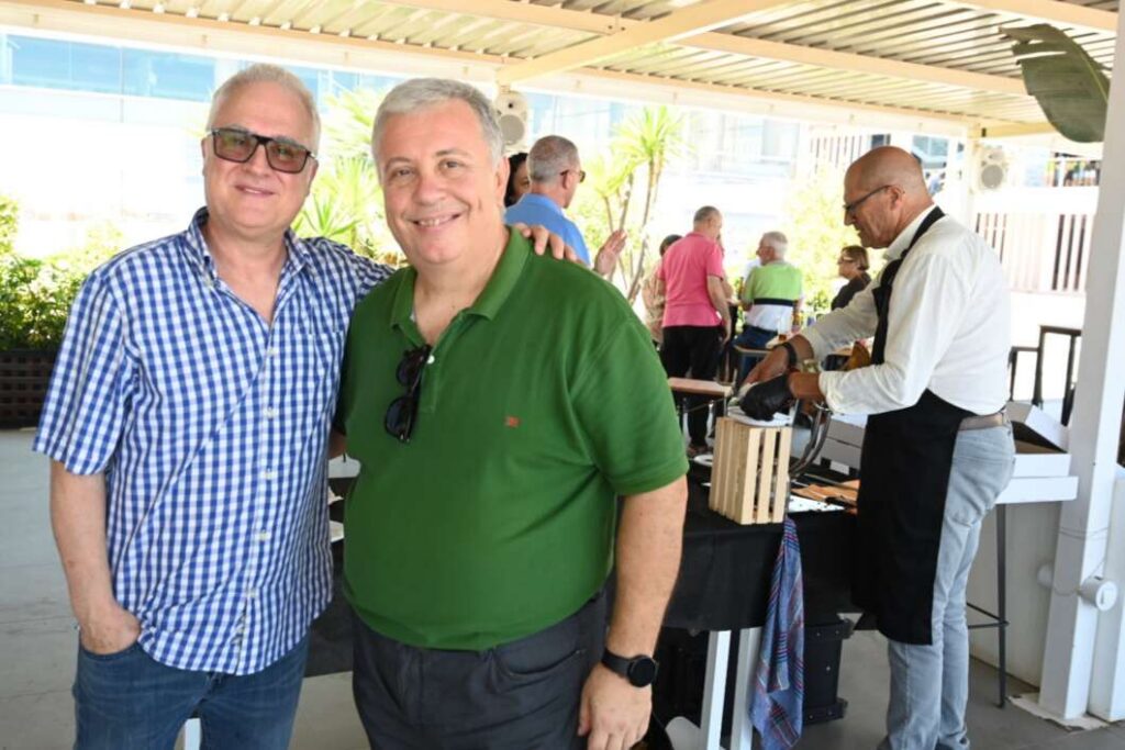 Two smiling men posing at an outdoor social event; one in a blue check shirt, the other in a green polo with sunglasses hanging from his chest.