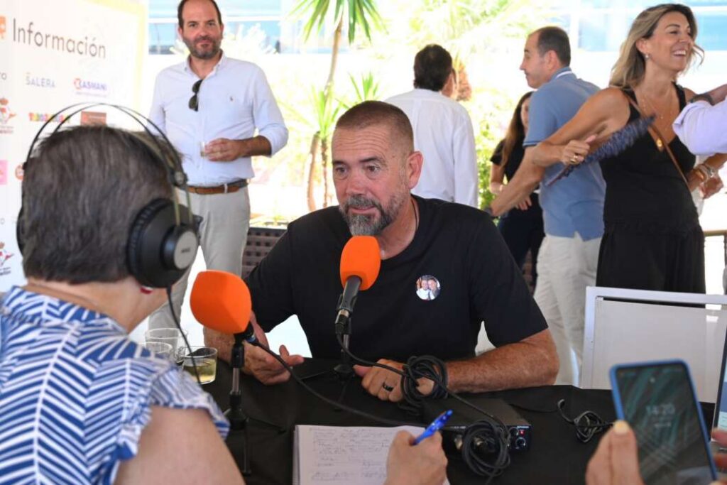Man with short hair and beard speaking into an orange microphone during a live podcast interview at a table; background shows attendees socializing outdoors under bright light.