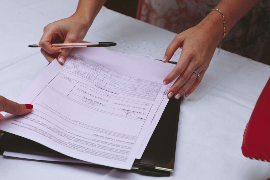 Hands arrange and review forms on a clipboard with a pen nearby on a table, a wedding ring visible on one finger.