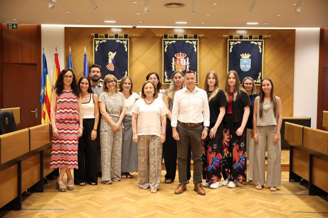 Group of fourteen adults posing for a formal group photo in a council chamber with flags and crest banners behind them.