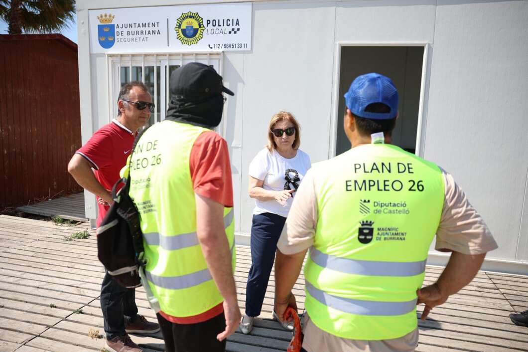 People in high-visibility vests labeled Plan de Empleo 26 talk with a woman wearing sunglasses outside a municipal building with a Policía Local sign.