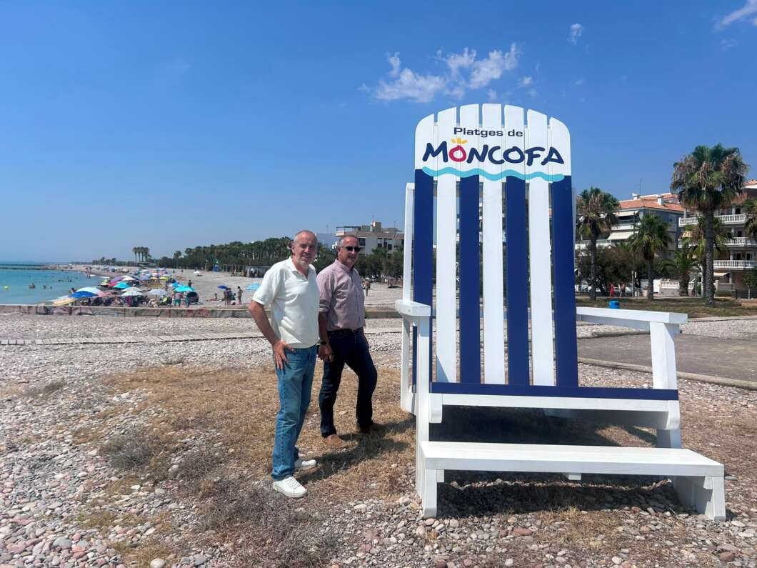 Two men pose on a pebbly beach beside a giant white-and-navy striped beach chair with a 'Platges de MONCOFA' sign on top.