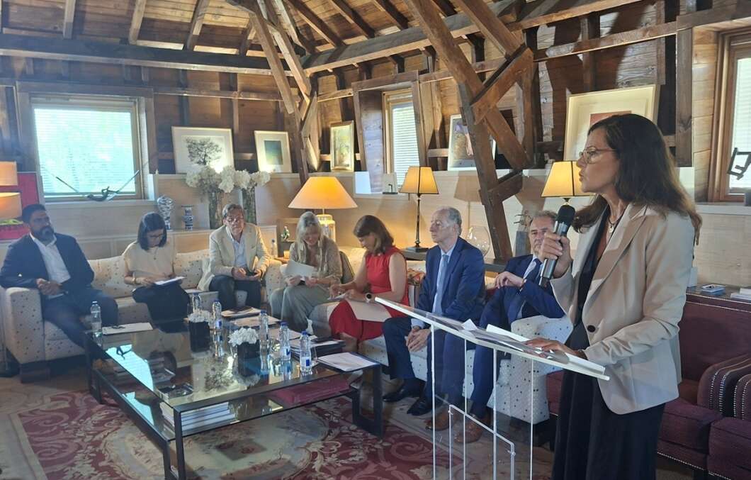 A woman speaks into a microphone at a lectern while a panel sits on a couch in a warm, wood-beamed room during a discussion event.