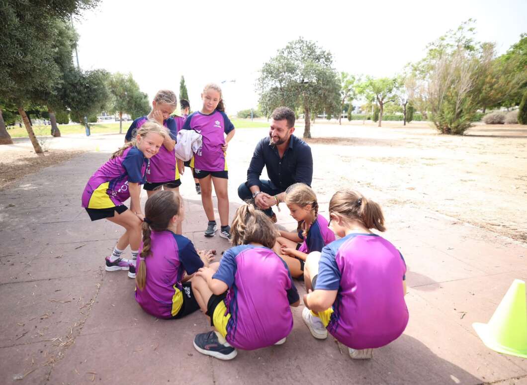 Group of young girls in purple and yellow sports uniforms sit in a circle on a park path, listening to a man kneeling and talking to them on a sunny day.