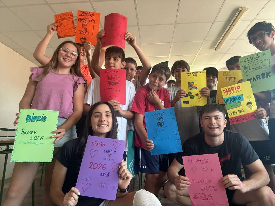 Group of smiling students in a classroom holding colorful handmade notebooks labeled 'Diario 2026' and 'Campus de Verano'.
