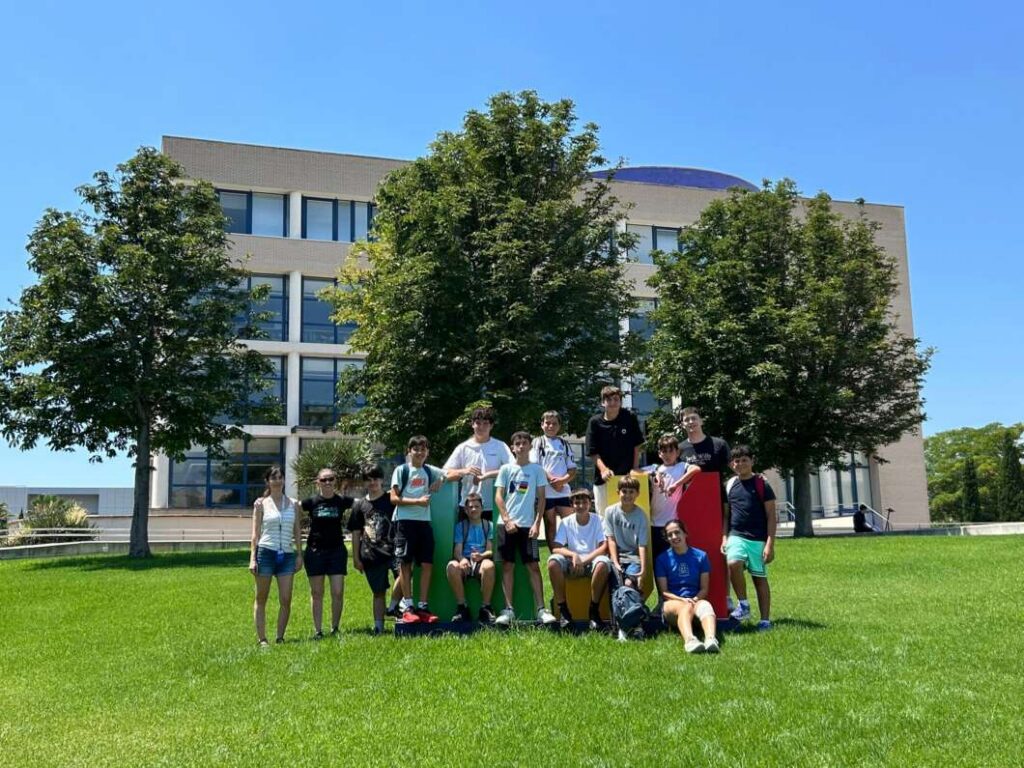 Group of diverse students posing for a photo on a sunny campus lawn in front of a beige multi-story building and trees.
