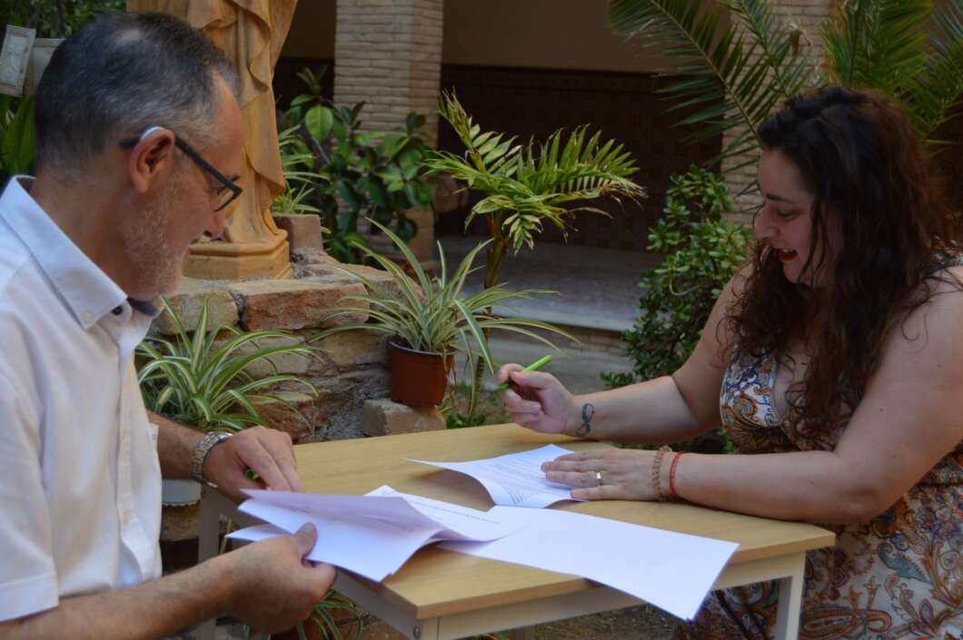 Two adults sign and sort papers at an outdoor table surrounded by plants and stone features.
