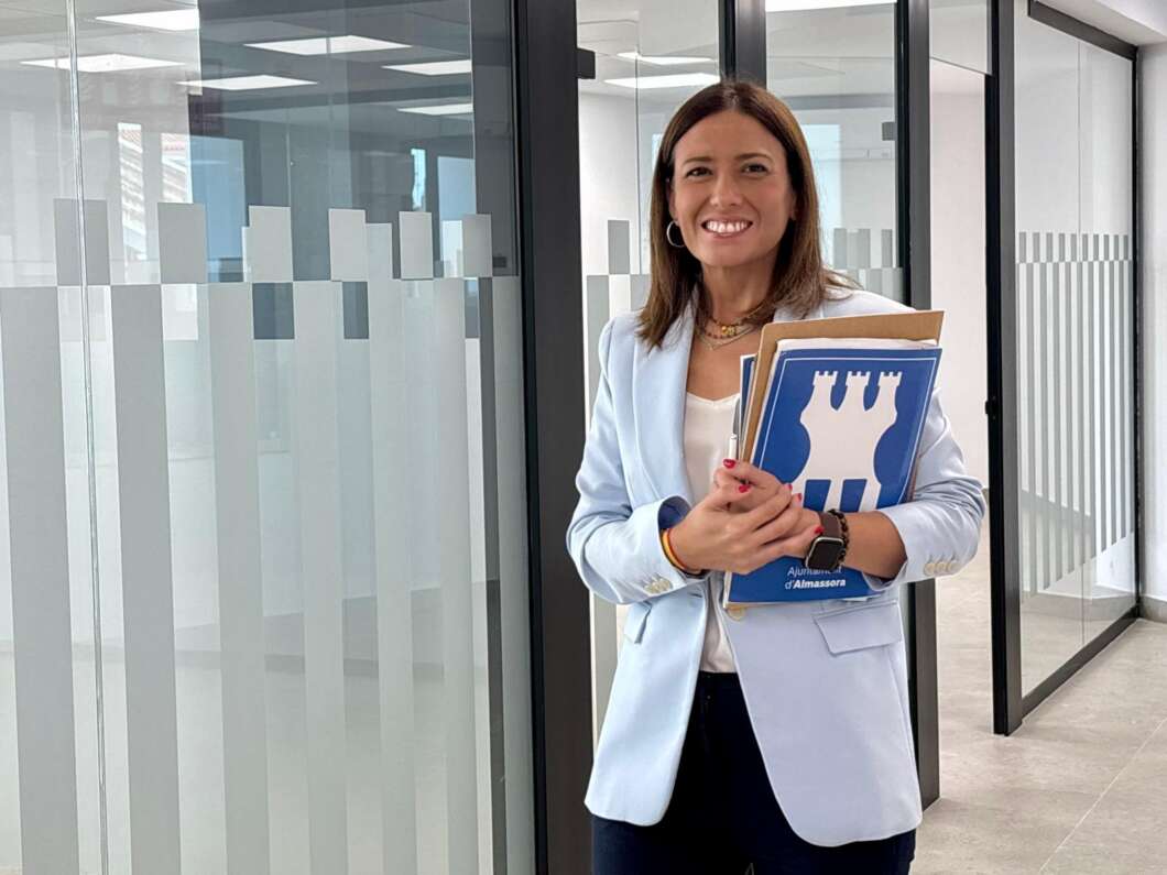 Smiling woman in a light blue blazer holding folders in a modern glass-office hallway.
