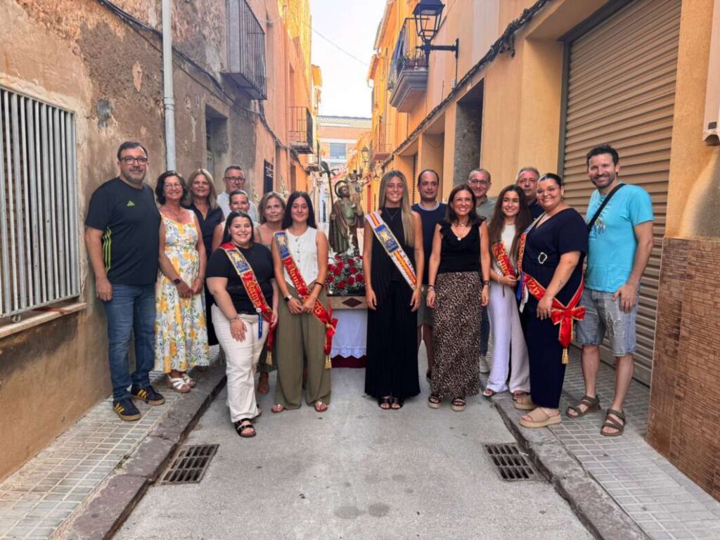 Group of people posing in a sunlit narrow street with a religious statue on a decorated table, some wearing sashes and festive attire.