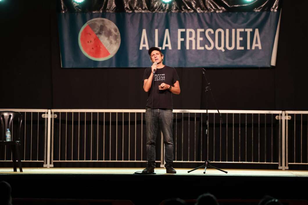Male performer on stage holding a microphone with a 'A LA FRESQUETA' banner featuring a watermelon behind him.