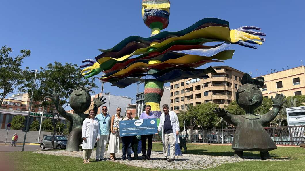 Group of eight people posing in a park in front of a tall multi-colored sculpture, holding a large check for 1,001 euros.