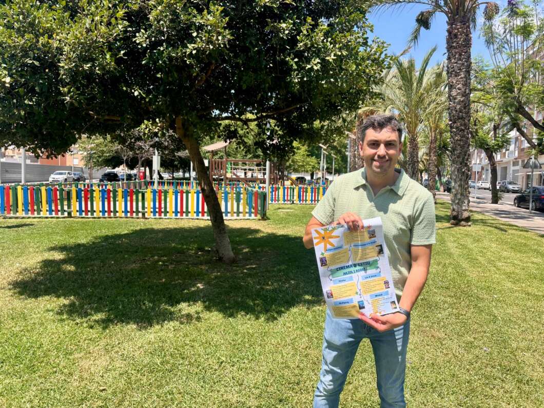 Man in a light green polo stands on the grass in a sunny park, holding a printed flyer or poster as a colorful fence and palm trees tower behind him.