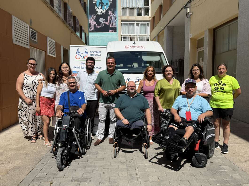 Group of mixed adults posing outdoors in front of a white van, two people in wheelchairs in the front row, smiling for the photo.