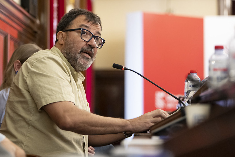 Bearded man with glasses speaking into a microphone at a podium during a panel or talk, beige shirt and audience in view