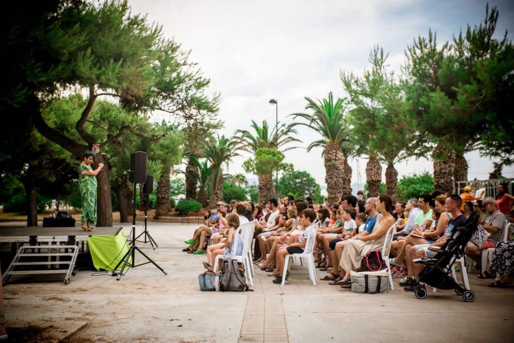 Speaker on a small stage addresses an audience seated in plastic chairs at an outdoor park with palm trees in the background.