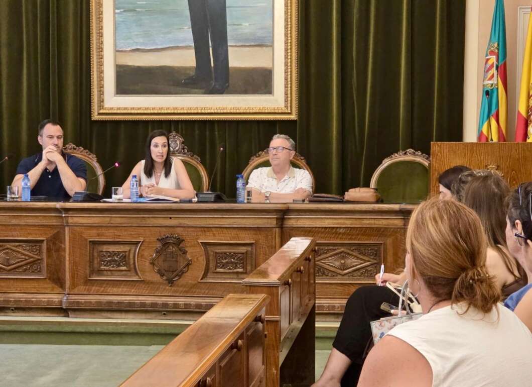 Panel of three people seated at an ornate wooden dais during a formal meeting, microphones and water bottles visible. Audience members take notes in the foreground.