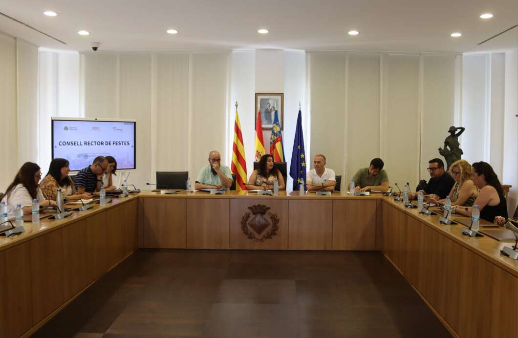 Council meeting in a boardroom with people seated around a U-shaped table; flags and a crest visible in the background, panel at the center.