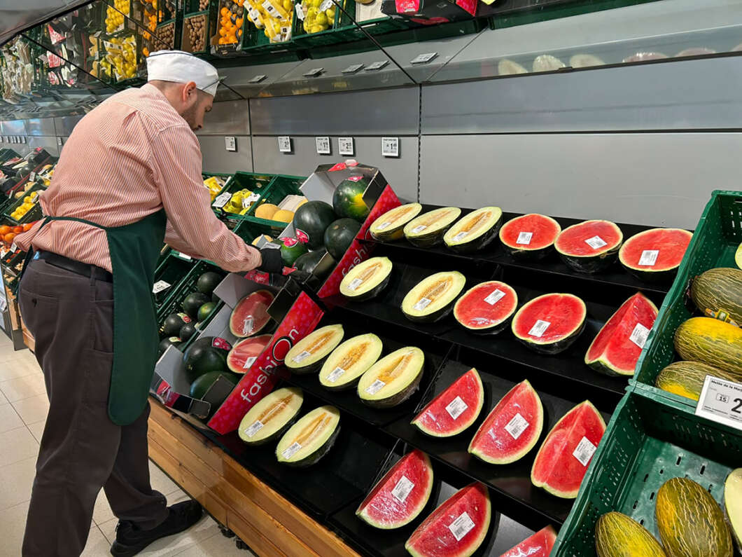 Male worker in a white hat and green apron arranges halved watermelons in a produce display at a supermarket.