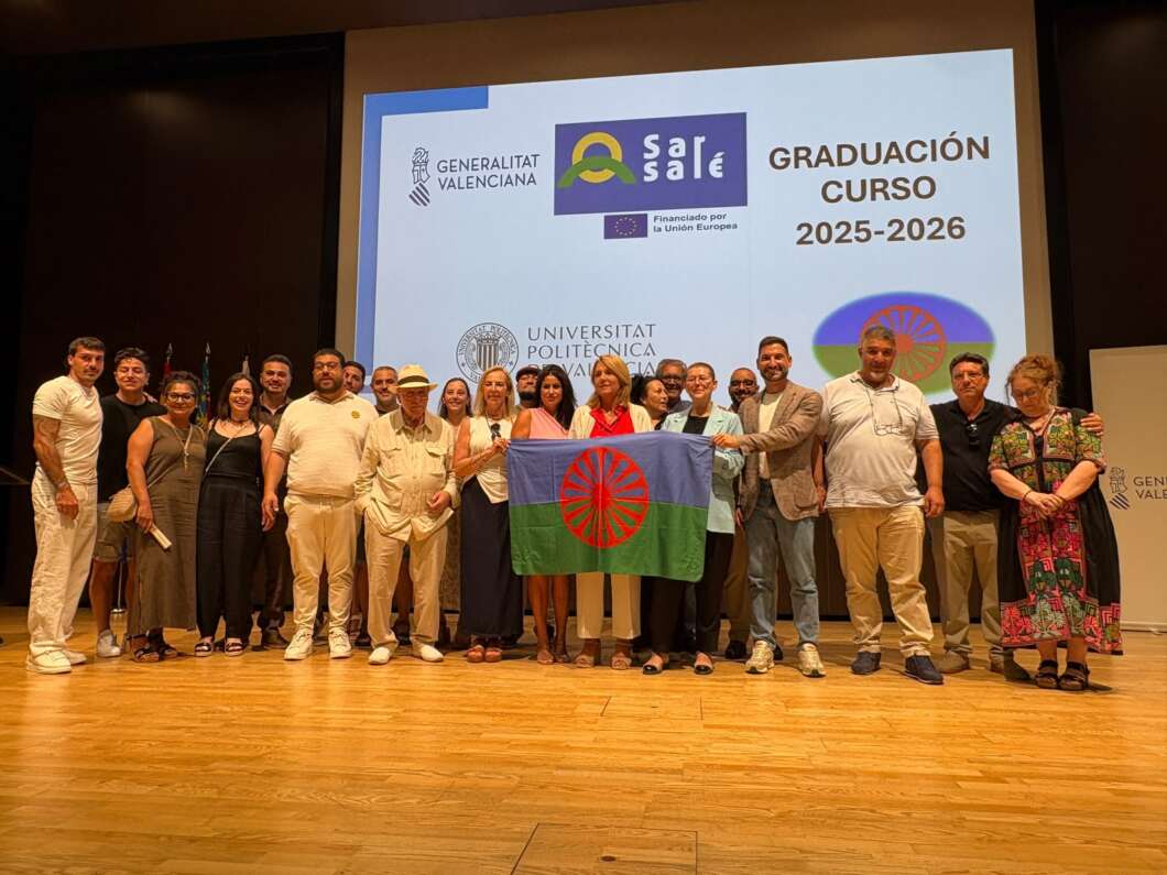 Diverse group of people on a stage posing for a graduation photo, holding a flag with a red circular emblem in the center.