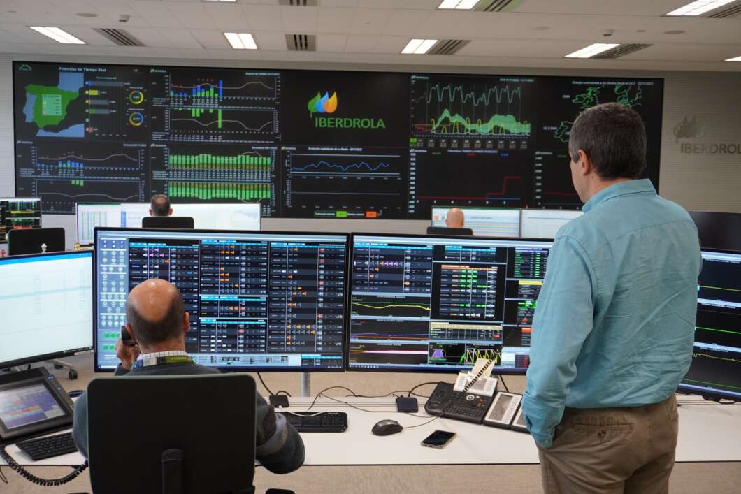 Two operators monitor multiple large screens displaying charts and data in a control room with Iberdrola branding.