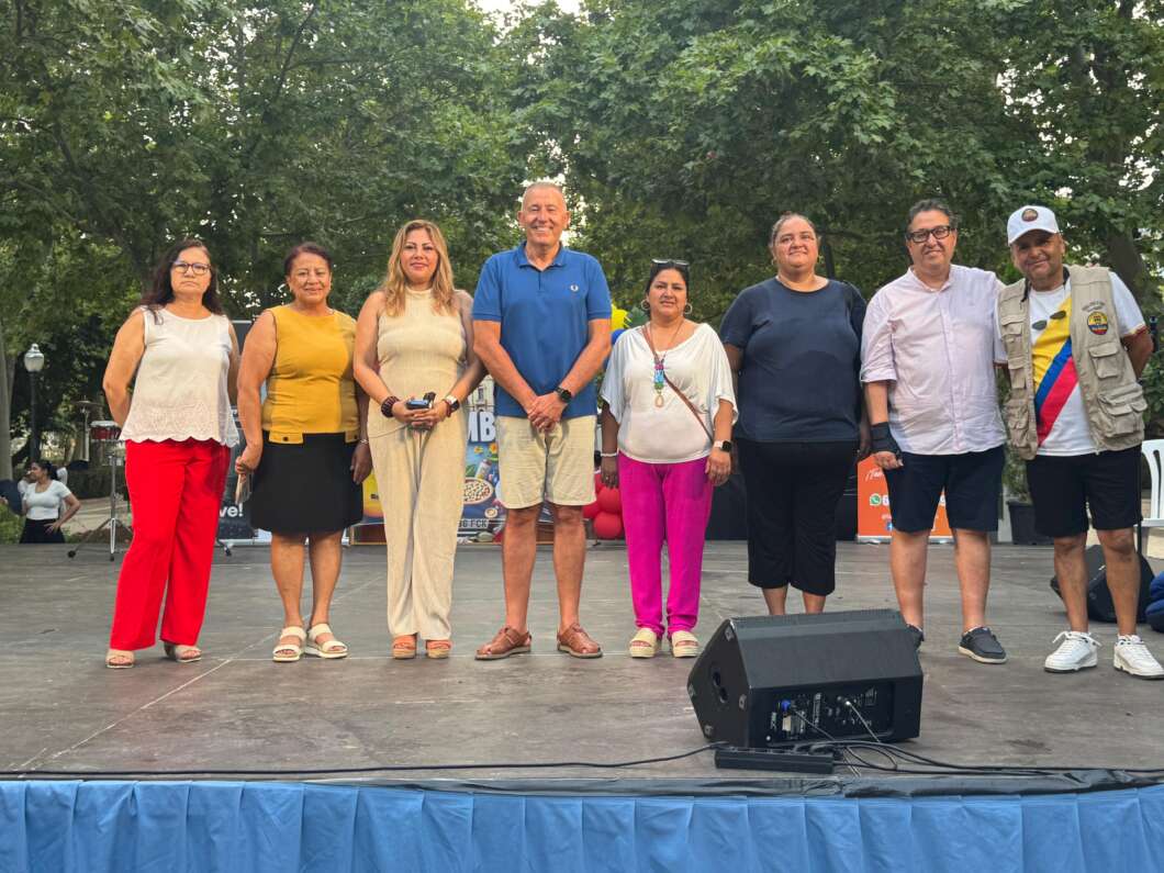 Nine adults stand in a line on an outdoor stage with trees in the background, posing for a group photo at a community event.