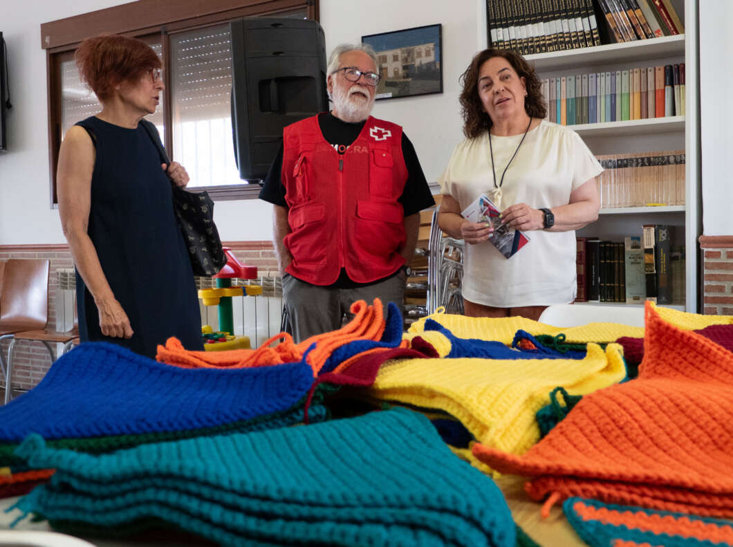 Three adults stand and talk in a room with bookshelves, colorful knitted blankets spread across a table in the foreground.
