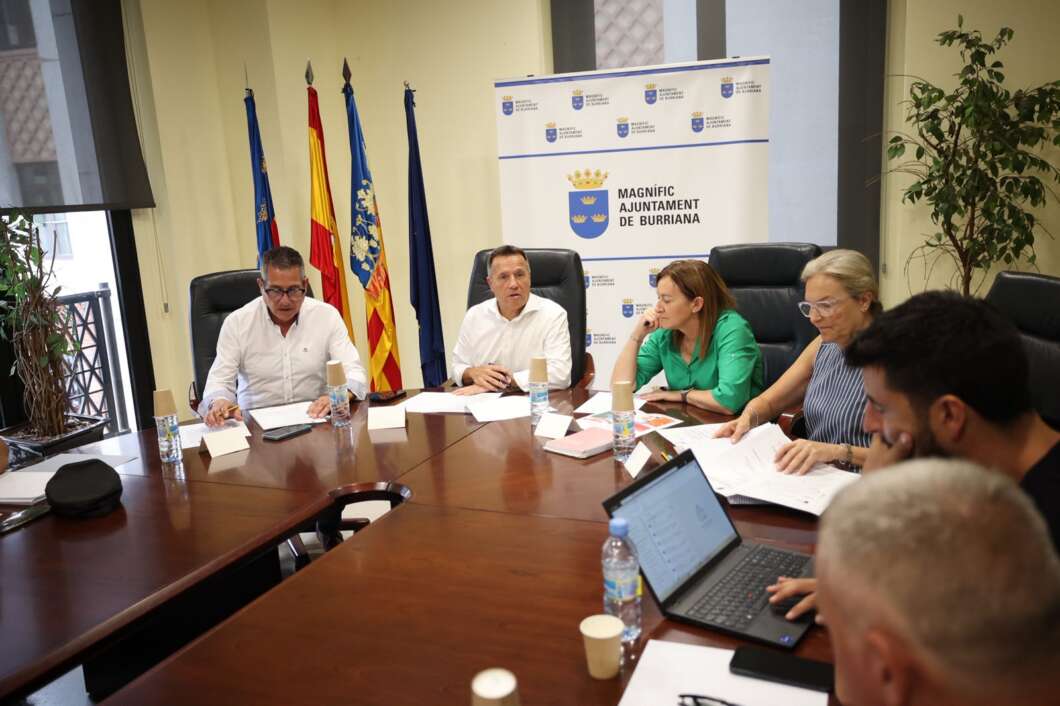 Group of people in a formal meeting around a wooden conference table with laptops, papers, and water bottles; banner and multiple flags visible in the background.