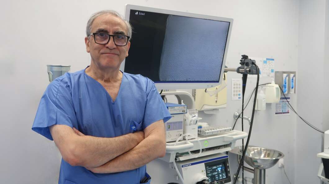Surgeon in blue scrubs with arms crossed, standing in a clinical operating room beside monitoring equipment and a medical cart.