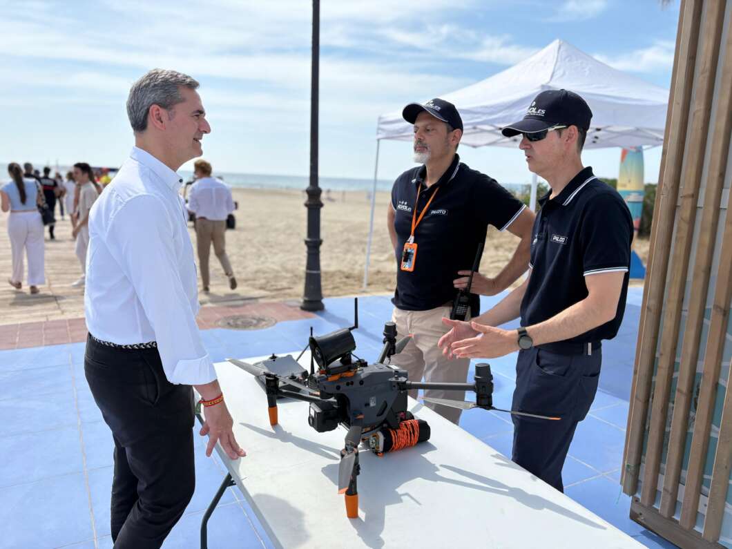 Man in a white shirt chats with two technicians beside a drone on a table at a beachイベント with the ocean visible in the background.”] ,